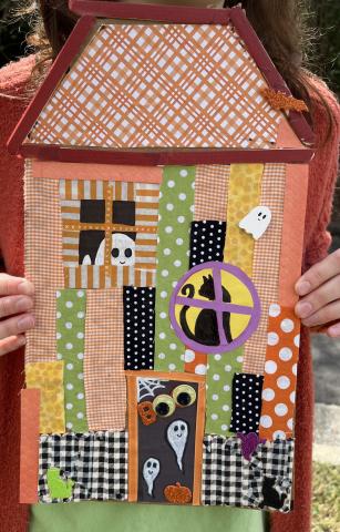 Pictured is a demo craft of a haunted house made with different craft materials such as washi tape, paint, craft sticks, fabric, and stickers.